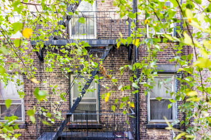 Brick apartment building exterior in Manhattan with fire escapes, windows, green tree leaves