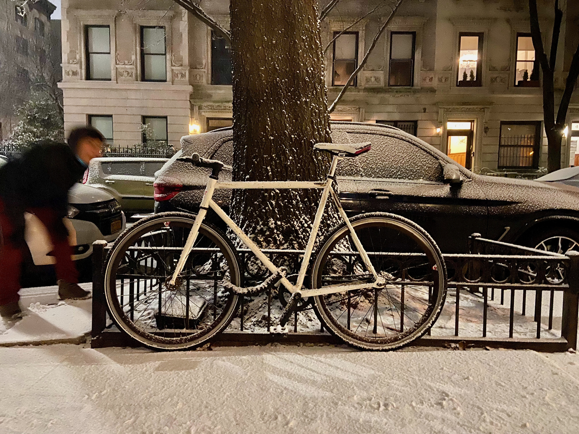 January 16, 2022 : A white bike chained to a fence in front of a tree with a woman bending next to it on a sidewalk in a residential neighborhood in Harlem, New York City, during a snow storm in the evening