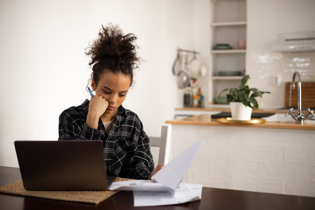 Young Black woman looking at documents at home, using a laptop