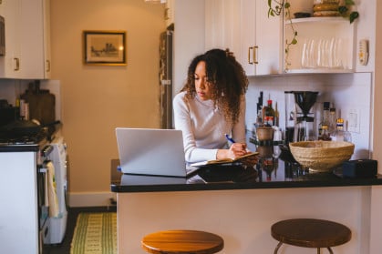 A young lady studying at the kitchen counter, utilizing a laptop and notepad.