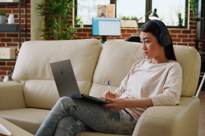 Focused working woman wearing wireless headphones taking notes while doing remote work at home sitting on sofa. Serious looking student attending online class on modern laptop.