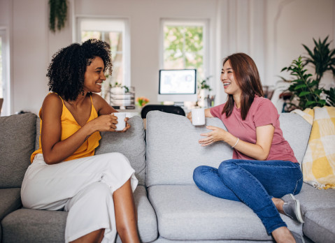 Two women sitting on sofa at home, they are drinking coffee and talking.