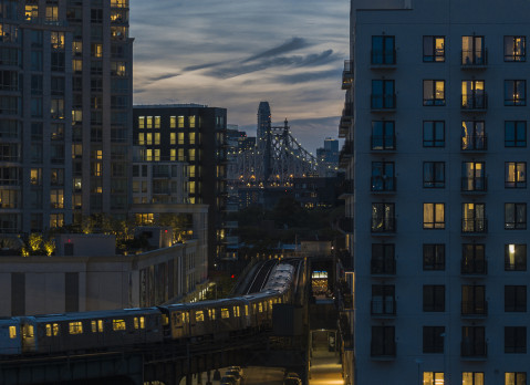 An elevated subway train riding on the line in Long Island City, Queens with Queensboro Bridge behind.