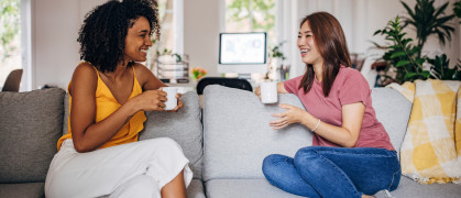 Two women sitting on sofa at home, they are drinking coffee and talking.