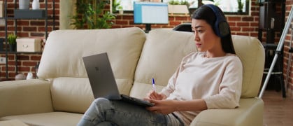 Focused working woman wearing wireless headphones taking notes while doing remote work at home sitting on sofa. Serious looking student attending online class on modern laptop.